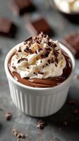 Creamy chocolate mousse topped with whipped cream and chocolate shavings served in a white ramekin on a dark countertop photo