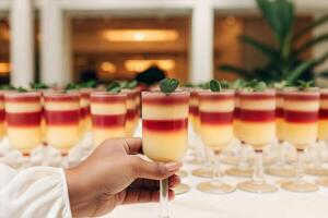 A photo of a sophisticated cocktail event with a hand holding a cocktail glass with a layered drink and a green garnish. Organized preparation for crowded party.