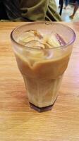 This drink is called palm sugar milk coffee, served in a glass and placed on a wooden table. The photo was taken from the top corner.