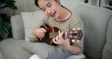 Handheld high angle view shot, Relaxed woman sitting on a sofa enjoying playing the ukulele in a cozy room at home. Practice and dedication to favorite musical instrument. video