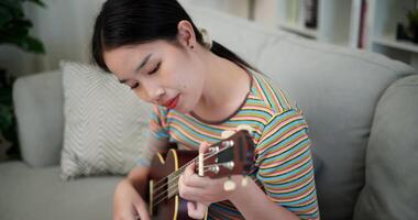 Handheld high angle view shot, Happy Asian woman playing ukulele on sofa in relaxing and warm atmosphere in living room at home. Enjoying the moment video