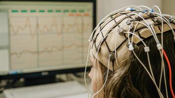 A detailed view of an EEG cap on a human head with electrodes attached to the scalp and data streaming on a nearby monitor visually representing the bridge between neural signals. photo