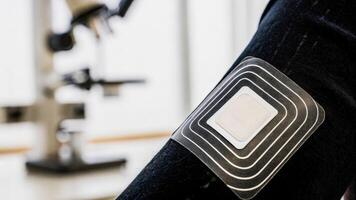 A closeup of a wearable patch adhered to skin showcasing its elegant design and tiny sensors with a softfocus background of laboratory equipment indicating advanced bioengineering.. photo