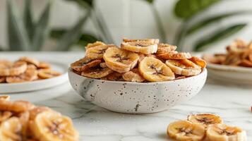 A bowl of dried banana slices on a marble surface, surrounded by greenery. photo