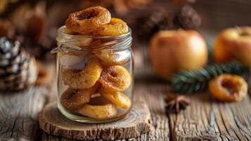 A jar filled with dried apple rings, surrounded by pine cones and apples on a wooden surface. photo