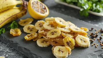 A display of dried banana slices alongside fresh bananas and lemon on a slate surface. photo
