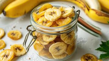A jar filled with dried banana slices, surrounded by fresh bananas on a white surface. photo