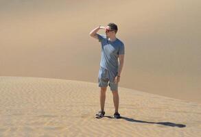 A man in a desert looking at view. A young man wearing a gray t-shirt and red sunglasses standing at the sand barchan background. Dune in Maspalomas, Gran Canaria photo