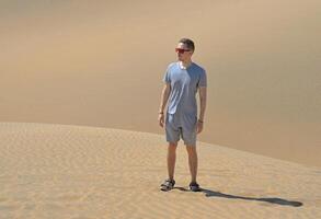 A man in a desert. A young man wearing a gray t-shirt and red sunglasses standing at the sand barchan background. Dune in Maspalomas, Gran Canaria photo