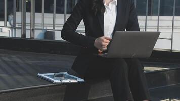 A woman in a business suit sitting on a ledge with a laptop video