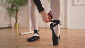 Ballerina puts on pointe shoes on leg in dance class. Classical ballet dancer woman in dance studio. Ballerina putting on tying classical pointe shoes for dance training. Ballet school concept video