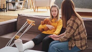 Girl with broken leg in cast sits with mother on couch at home. Sad little kid hugging teddy bear recovering from broken leg. Girl using cast crutches for quick rehabilitation. Mom supporting daughter video