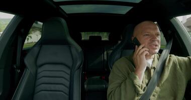 A bald man sitting in the back seat of a car talking on a cell phone video