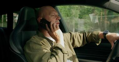 A bald man sitting in the driver's seat of a car talking on a cell phone video