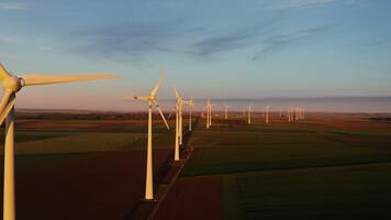 A lot of rotating wind turbines against the background of multi-colored fields in the red rays of the setting sun, a bird's eye view from a drone. video