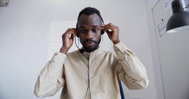 Preto cara com uma barba dentro uma camisa, com uma curto afro Penteado com a brinco dentro dele orelha desgasta fones de ouvido com uma microfone sorridente fala e alterar dentro a face fechar-se. video