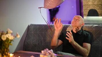 Bald man catching heart shaped balloon while seated on restaurant couch, expressing whimsical romantic moment in soft, intimate lighting Valentine's Day video