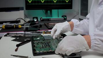 A technician wearing white gloves and a lab coat is carefully repairing a laptop motherboard on a workbench. The background features electronic tools and a monitor displaying a waveform. video