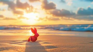 A creative Easter concept featuring a red paper rabbit standing on the beach with the ocean and a golden sunset in the background. photo
