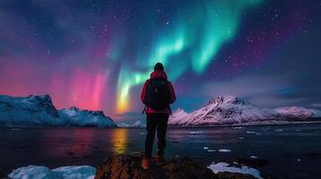 Person with backpack enjoying stunning Northern Lights display over snow covered mountains and calm water at night photo