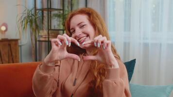 Happy young Caucasian woman sitting on couch makes symbol of love, showing heart sign to camera video