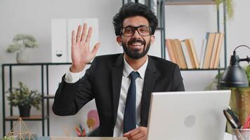Businessman working on laptop smiling friendly at camera and waving hands gesturing hello at office video