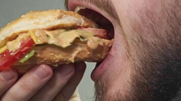 Young man with beard bites into juicy burger with high-calorie sauce, enjoying its taste as he savors it, licking his lips to remove last traces of delicious sauce. Close up. Portrait. video