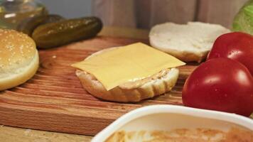 Cook prepares homemade burger, adding square piece of melted cheddar cheese and chicken patty to bun. On table, there are traditional ingredients for an American burger, such as pickles and tomatoes. video