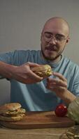 Bald man with beard is excited about trying delicious homemade hamburger. After taking bite, he enjoys mouth-watering flavor of food. Vertical. Portrait. video
