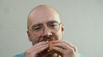 Hungry bald man wearing glasses is eating juicy hamburger with great appetite and enthusiasm, looking directly into camera and smiling on white background. Portrait video