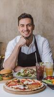 A man in an apron is smiling while sitting at a table with pizza, salad and drinks photo
