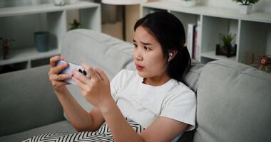 Handheld high angle view shot, Young asian woman wearing wireless headphones sits on a sofa with eyes wide open and mouth slightly open, indicating shock or surprise while looks at mobile phone video
