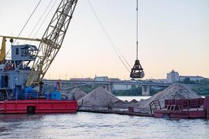 River port. Port cranes on the river bank On a sunny, beautiful day Extraction and transportation of sand and other goods. The embankment of the river port, a barge at the mooring wall. photo