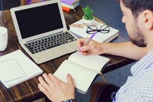 Young man working at desk, writing notes in a notebook while using a laptop in a study environment photo