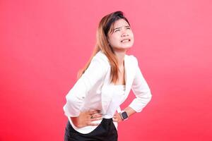 A woman wearing a white jacket and black pants holds her stomach with both hands and shows a facial expression of pain isolated on a red background photo