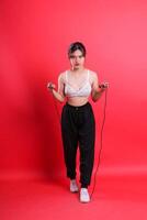 A person in a white top and black pants is holding a jump rope in front of them, isolated on a red background. photo