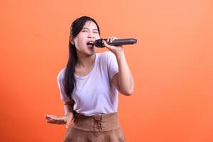 An enthusiastic young woman performs a song with a microphone, expressing pure joy and energy, isolated on orange background photo