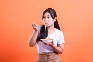 A young Asian woman is happily eating a bowl of noodles with a fork, she seems to really enjoy it, isolated on orange background photo