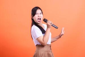 A young woman with a microphone singing passionately with her mouth open, hand raised and expressive face, isolated on orange background. photo