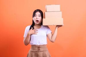 A young Asian woman is surprised while holding three stacked cardboard boxes in her hand, with an expression of shock. isolated on orange background photo
