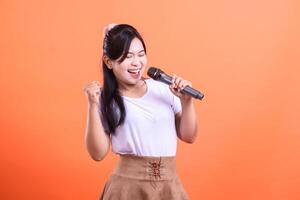 A young and excited woman holding a microphone with a happy expression, is giving a performance with full energy, isolated on orange background photo