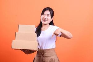 A happy woman holds a stack of cardboard boxes while pointing at them with her finger, she is smiling and cheerful. isolated on orange background photo