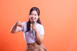 A young woman smiles broadly while singing into a microphone and pointing her finger down to the side, isolated on orange background photo