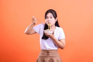 A young woman with a surprised expression, happily eats noodles with a fork from a white bowl. isolated on orange background photo