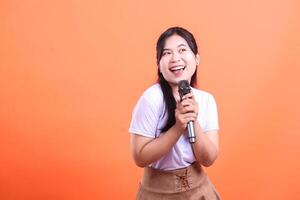 A joyous Asian woman smiles while holding a microphone, looking away, ready for her performance. isolated on orange background photo