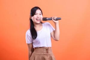 A smiling young woman holding a microphone, ready to sing a song or deliver a speech, captured mid-performance. isolated on orange background photo