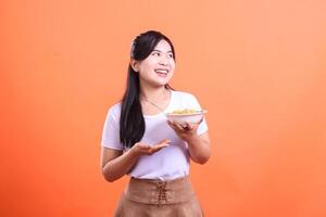 A joyful young woman is showing off a bowl of noodles, holding it in her hands. She is wearing a white t-shirt and brown skirt, isolated on orange background photo