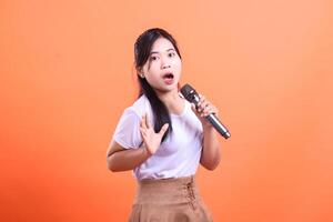 A young asian woman with open mouth, holding a microphone and gesturing with her hands, showing enthusiasm while singing, isolated on orange background. photo