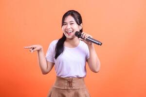 A young woman singing with a microphone and pointing with her left hand in a studio, she looks very happy and friendly. isolated on orange background photo