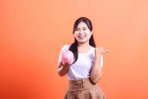 Happy asian woman with a piggy bank in her hands, pointing to the side. Financial literacy and future investments are her goal, isolated on orange background. photo